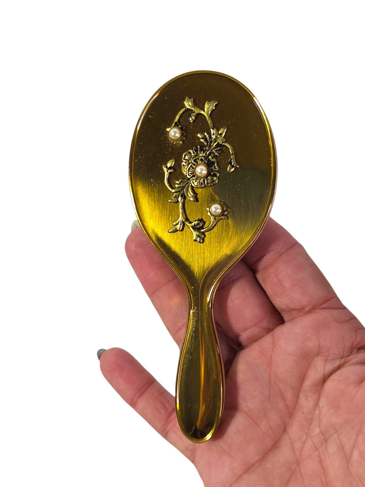 Back of a mini gold-tone hand mirror with an antiqued floral spray and three faux pearls, held in hand