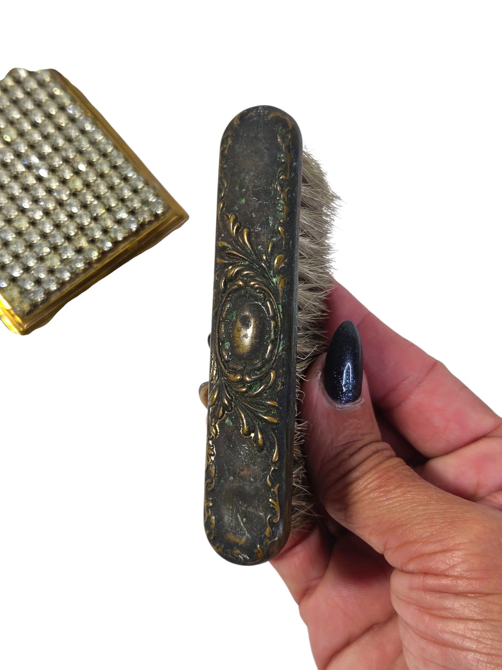 Small vintage clothes brush held in hand, repousse scroll metal back with dark patina and green verdigris, rhinestone compact beside it