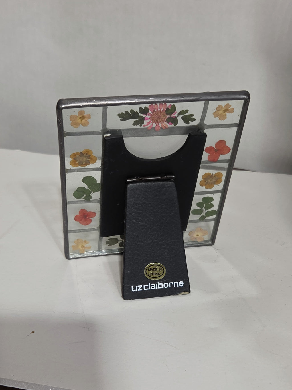 Back view of an Oddly Darling Vintage Liz Claiborne glass picture frame with real pressed flowers and a black stand.