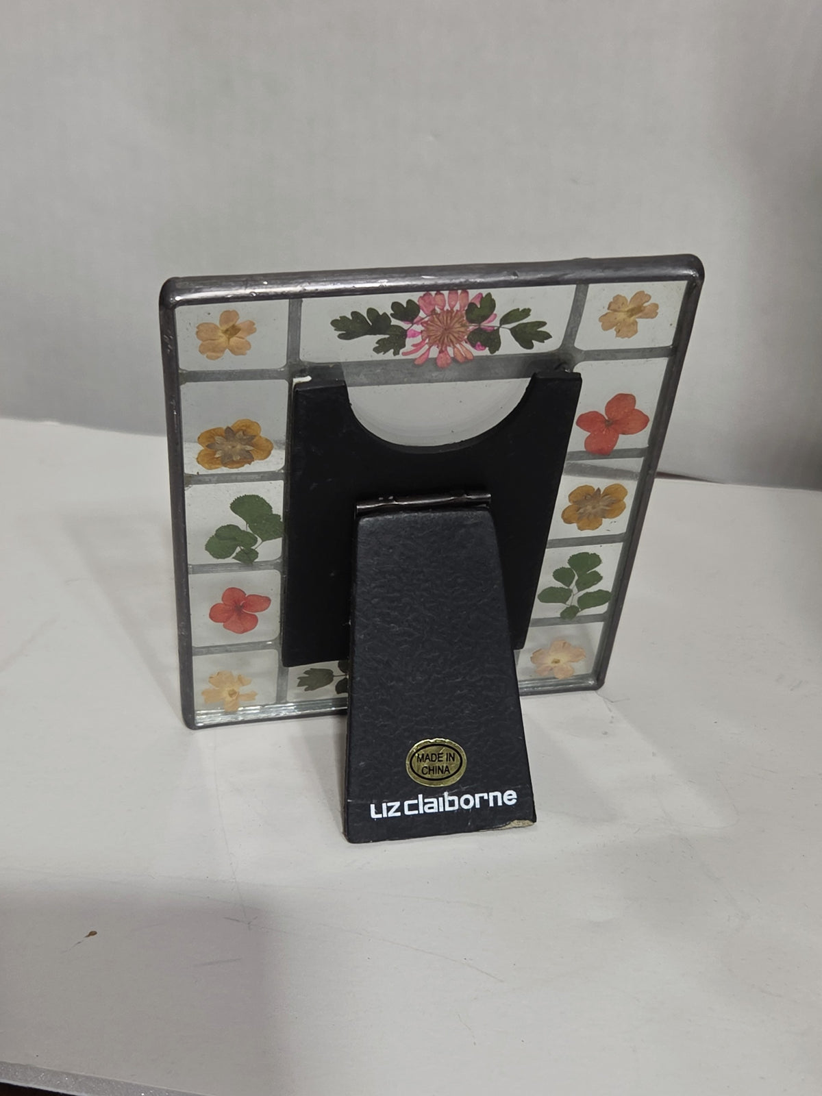 Back view of an Oddly Darling Vintage Liz Claiborne glass picture frame with real pressed flowers and a black stand.