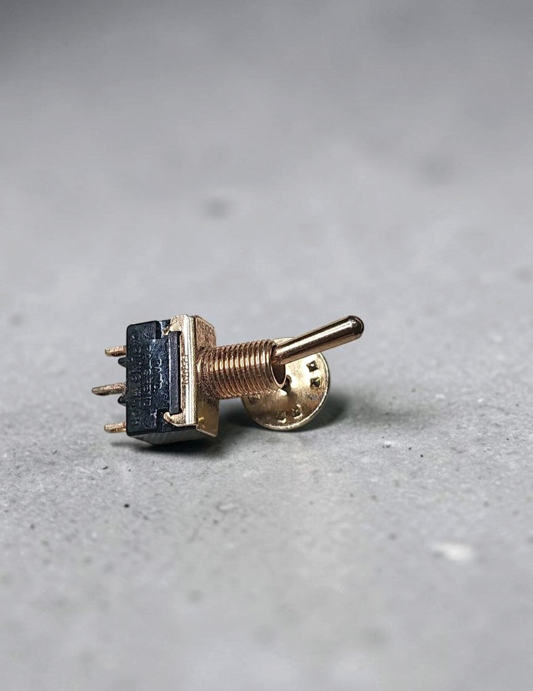 Oddly Darling Vintage gold-toned toggle switch lapel pin with a black housing on a grey textured surface