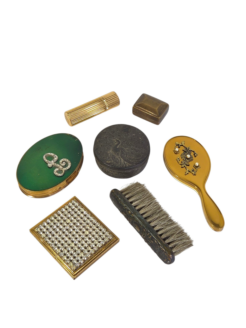 Vintage vanity lot arranged at an angle: green L compact, rhinestone compact, embossed peacock box, pearl-trimmed hand mirror, bristle brush, pill box and lipstick