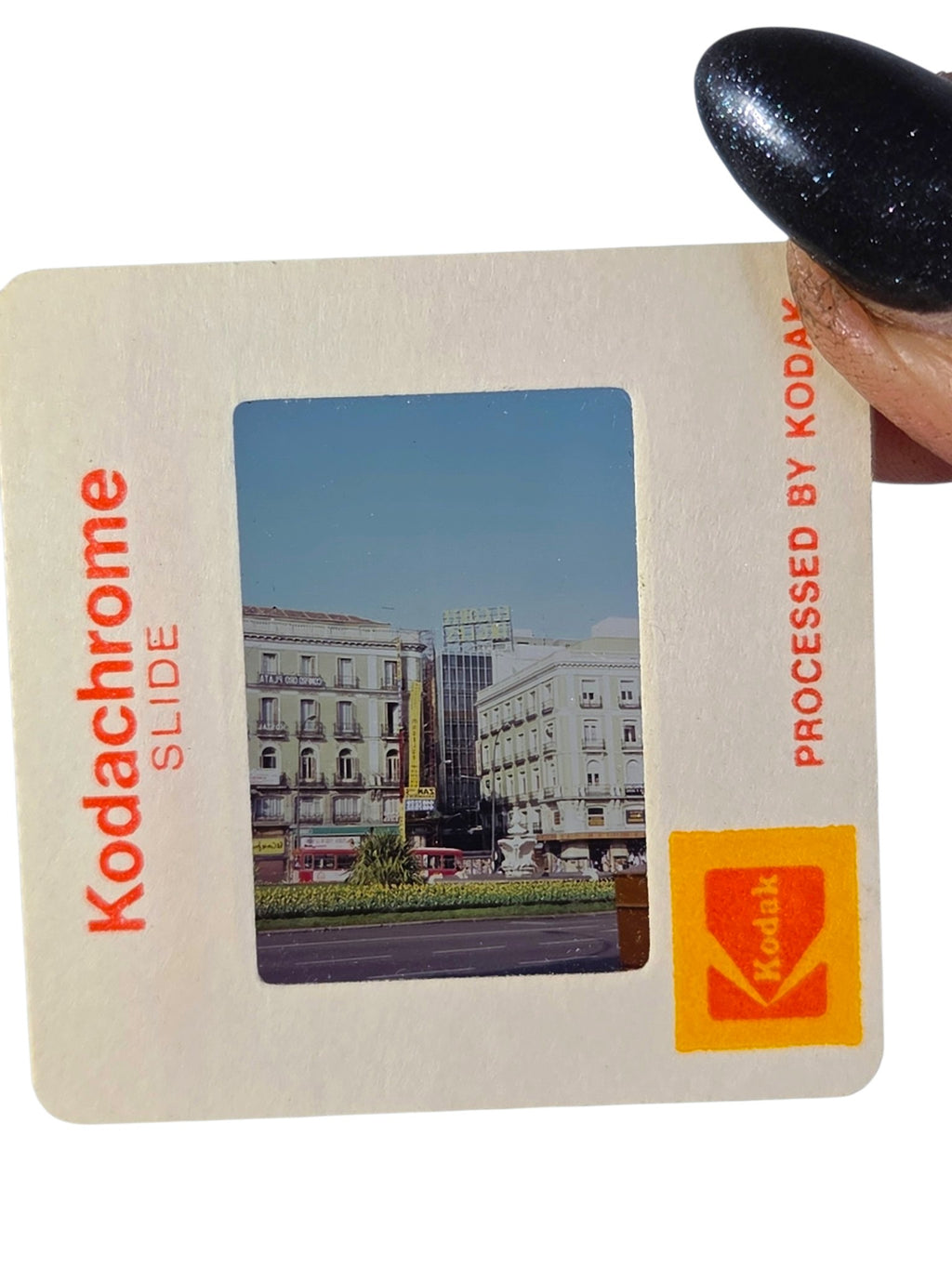 Kodachrome travel slide held in hand showing old city buildings, a fountain, flower beds and red buses under blue sky, likely Madrid